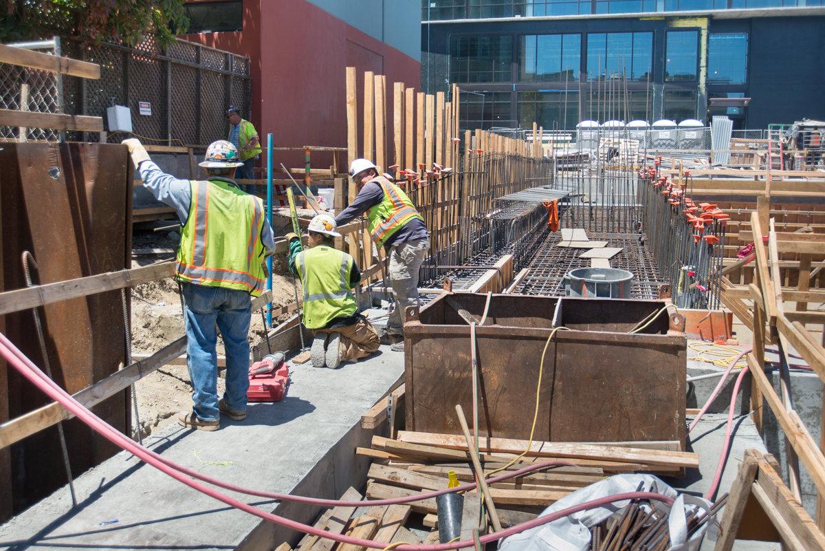 Construction workers in safety vests remove scaffolding from a transit station headhouse using a sledgehammer and scissor lift.