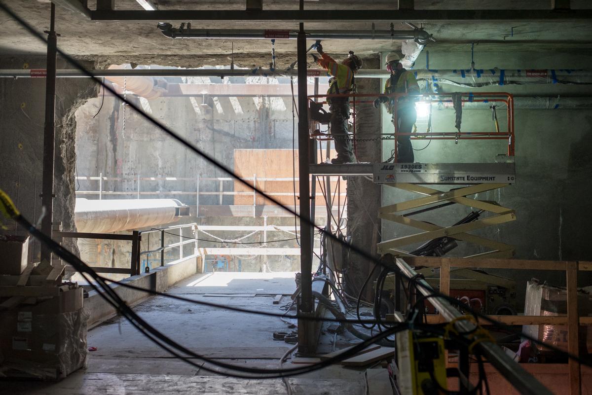 Construction workers install ceiling structural elements inside station box with plastic sheeting containment.