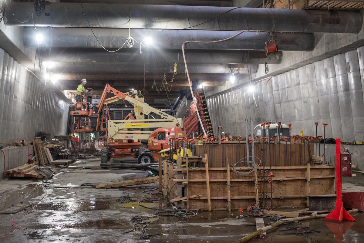 Workers remove concrete from temporary bracing inside underground transit station construction site using aerial lift and equipment.