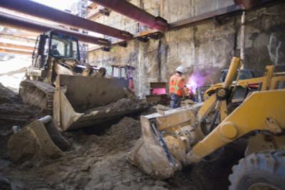 Construction workers operate heavy equipment inside a transit station headhouse during underground excavation and utility work.