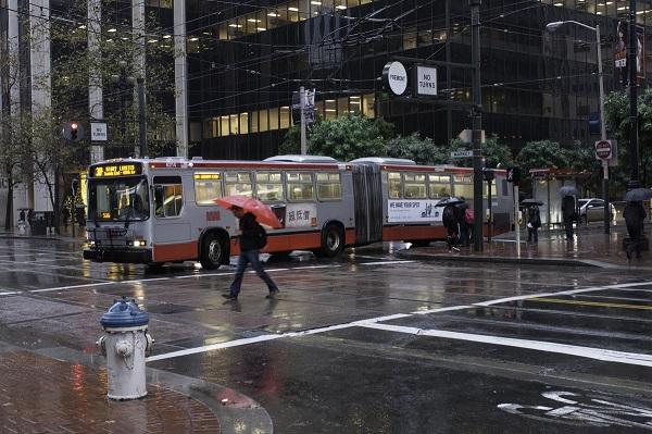 Red and white Muni bus stopped on wet street during rainy weather in urban downtown area.