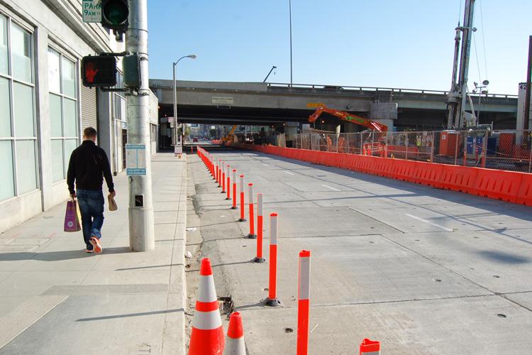 Pedestrian walking on sidewalk past red and white traffic barriers and construction equipment on 4th Street.