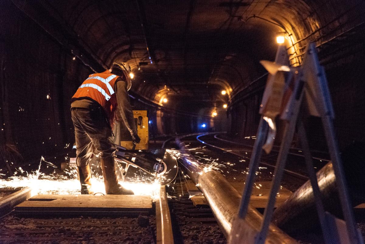 Worker in safety vest welding railroad track in tunnel at night with sparks flying.
