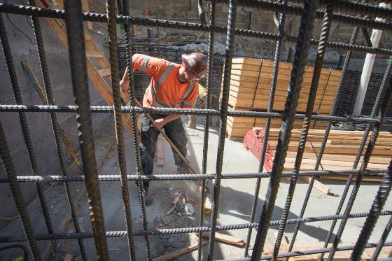 Worker in orange shirt installing rebar behind metal safety fencing at construction site.