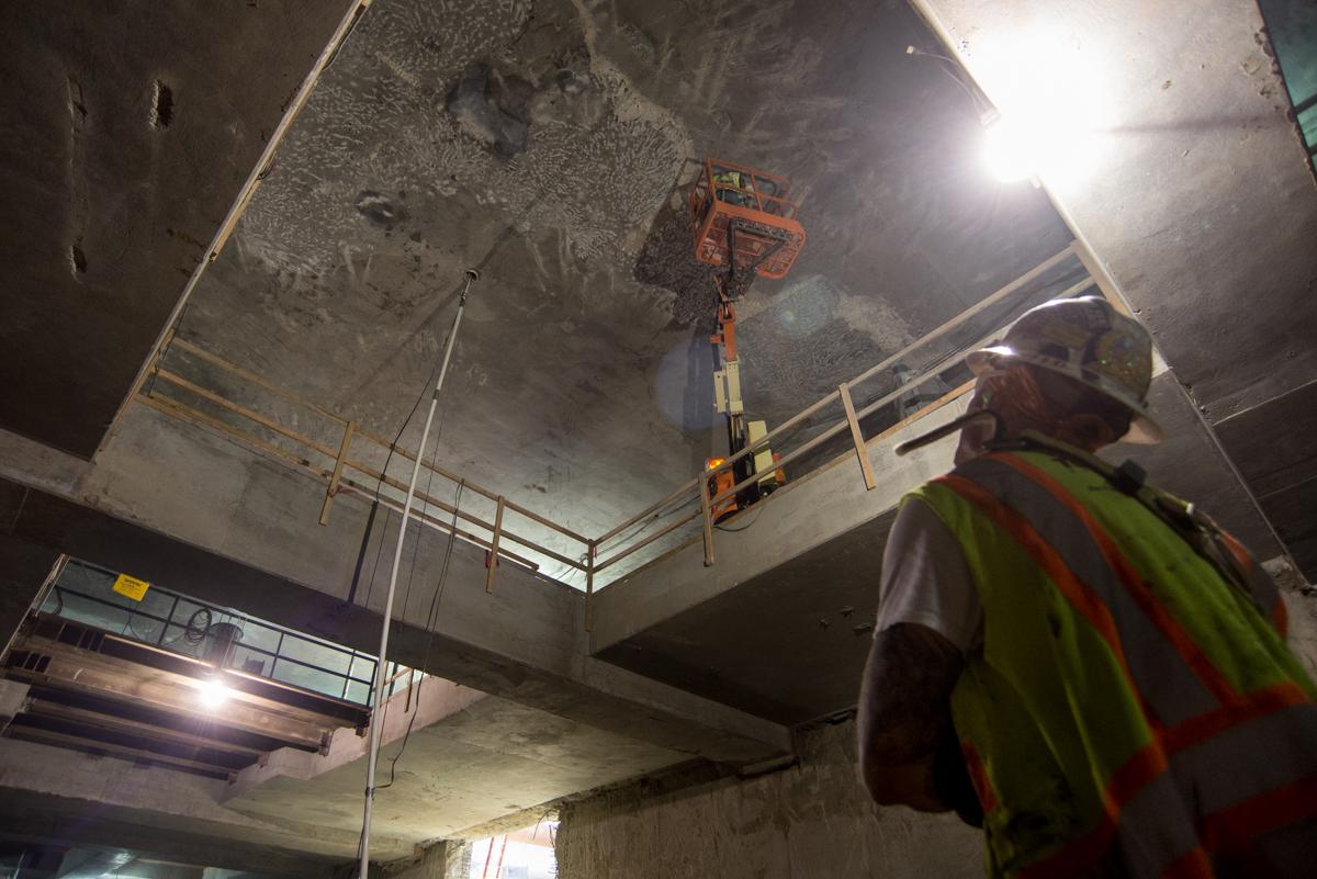 Worker in safety vest operates jackhammer on concrete ceiling of underground transit station mezzanine level.