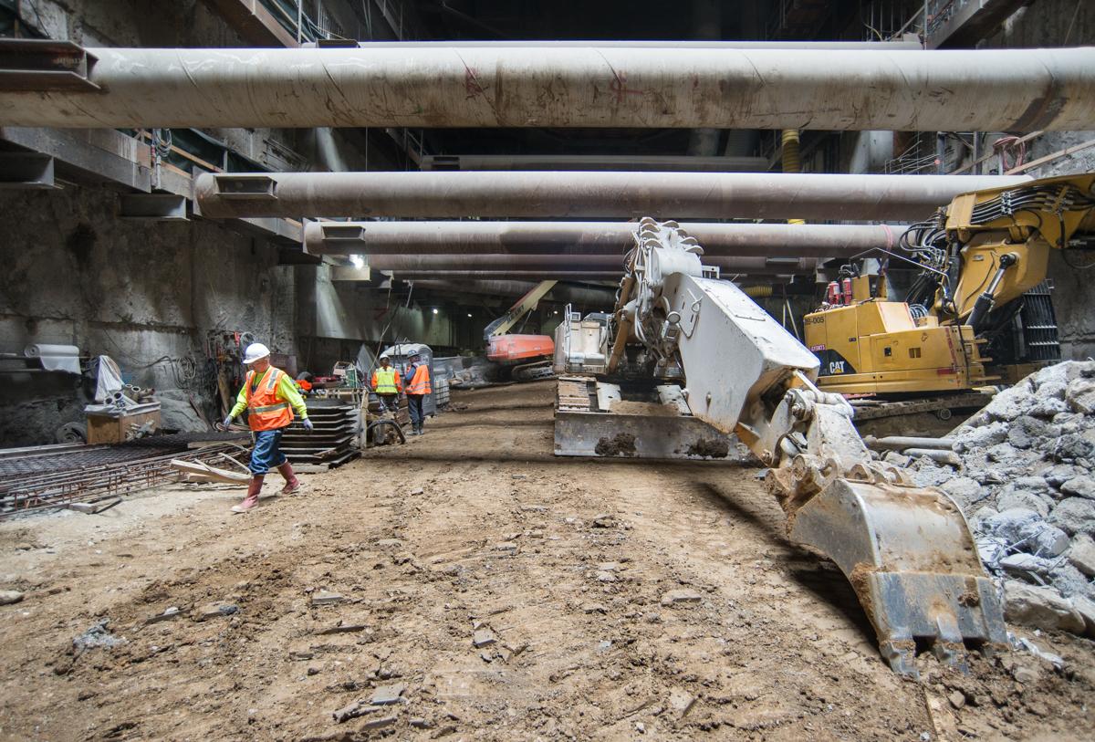 Construction workers and heavy equipment operating in underground transit tunnel excavation site beneath concrete structure.