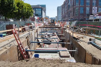 Construction excavation on 4th Street showing sewer line installation with equipment and urban buildings in background.