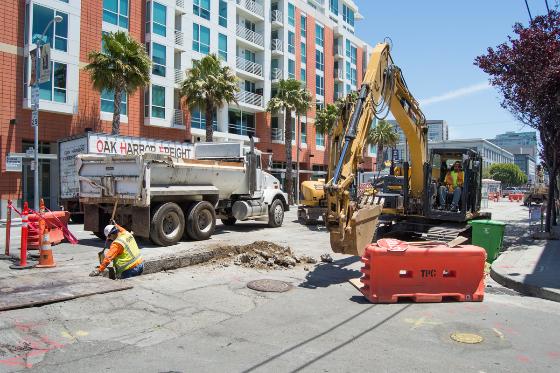 Construction excavator and dump truck on urban street with residential building in background.