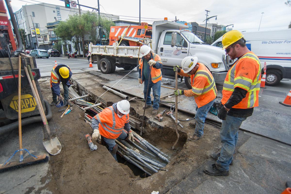 Construction workers in safety gear separate underground utilities in an open trench at an urban street intersection.