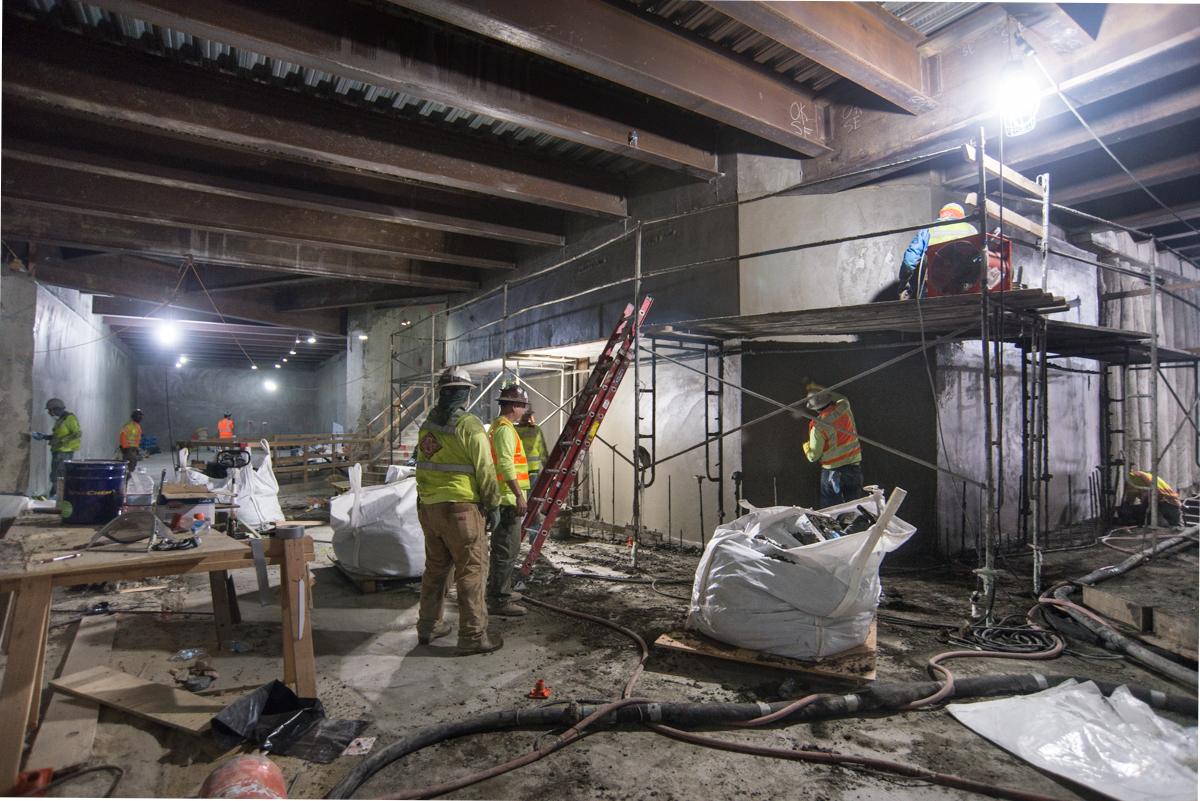 Construction workers in safety vests work on reinforced concrete walls in an underground transit tunnel with metal scaffolding and equipment.