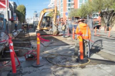 Construction crew operating excavator to backfill utility trench on urban street with red safety barriers.
