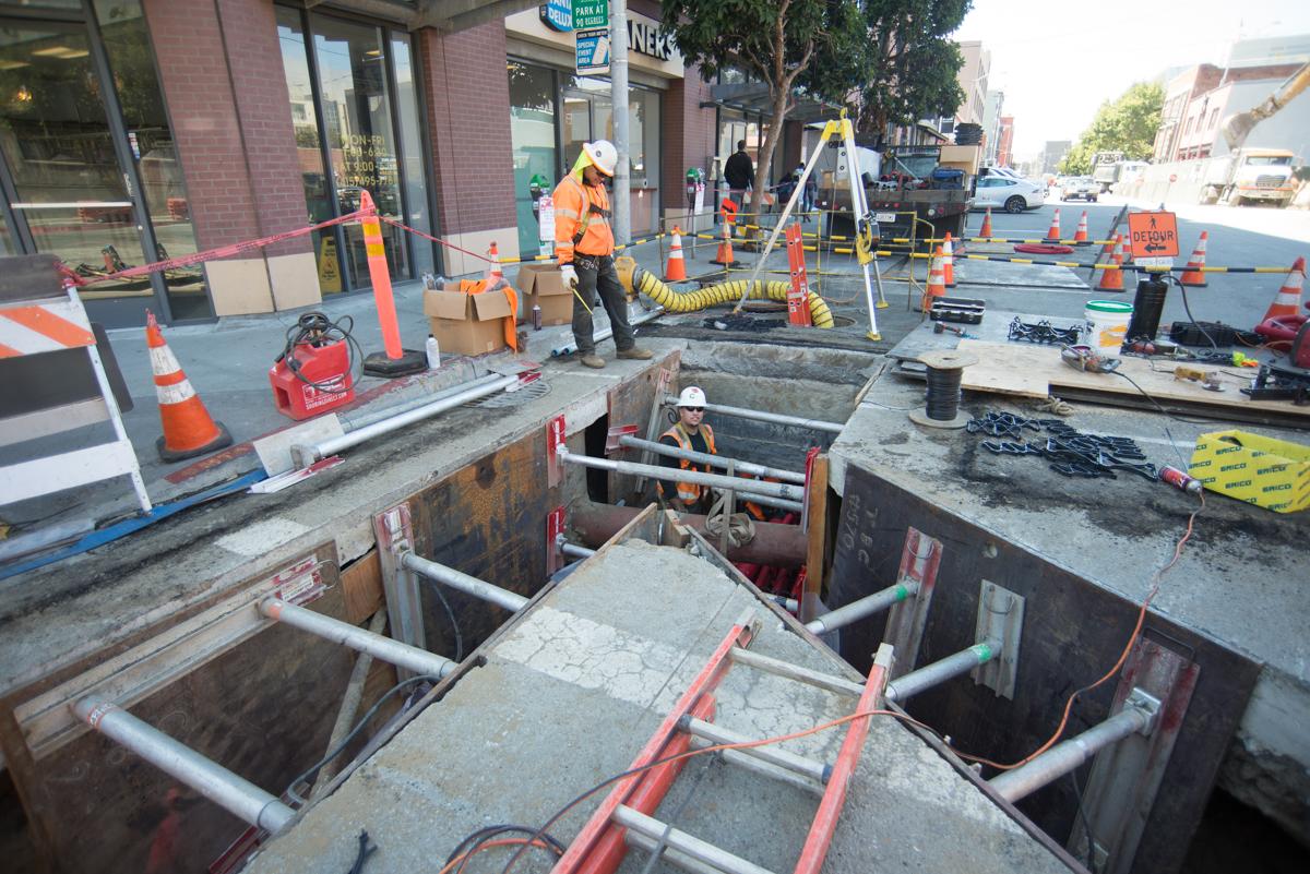 Construction workers in safety gear performing underground sewer work on a city street with excavation equipment and traffic control barriers.