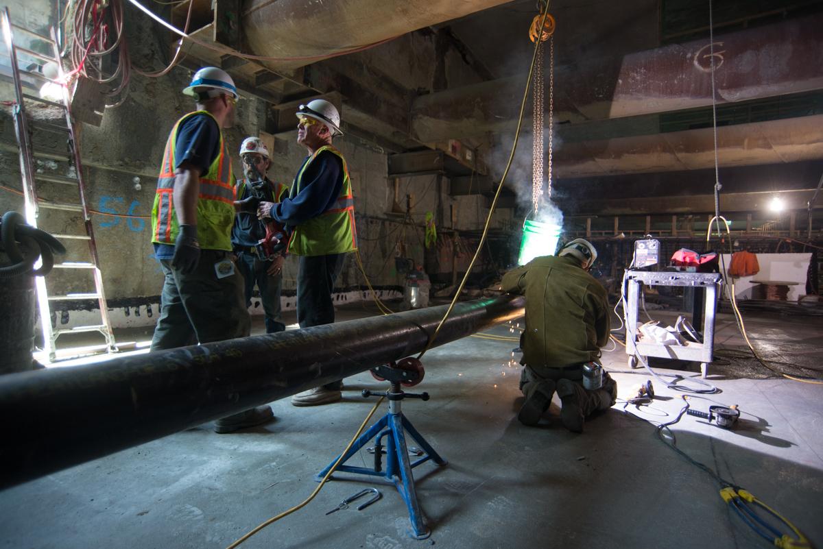 Workers in safety gear install steel pipe sections for track drainage during station construction.
