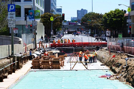 Construction site with excavation, roof deck installation, and workers in safety gear along urban street.