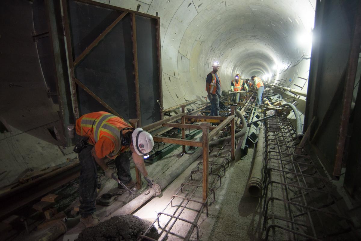 Workers in safety gear disassemble concrete pump hose inside illuminated transit tunnel under construction.