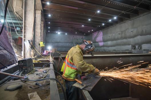 Worker in safety gear welding steel brackets in underground transit tunnel at night.
