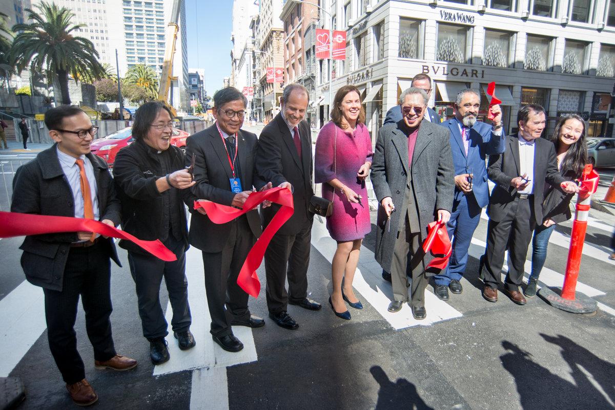 Group of officials and dignitaries cutting red ribbon at urban street ceremony with buildings and palm trees in background.