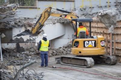 CAT excavator demolishing concrete section of Union Square Garage with worker in safety vest observing.