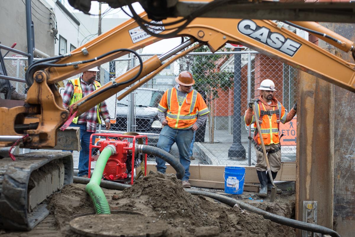 Construction crew performs utility work on 4th Street with excavator and water pipe equipment.