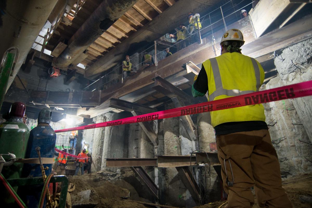 Worker in safety gear holding red barrier tape at underground construction site with steel structural work visible.