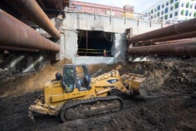 Bulldozer moving soil near underground station construction site with large pipes and building visible.