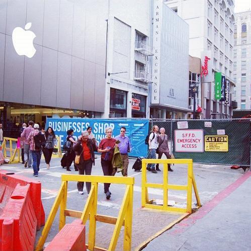 Construction site with yellow barriers and red fencing near Union Square, pedestrians walking through area with Apple Store visible.
