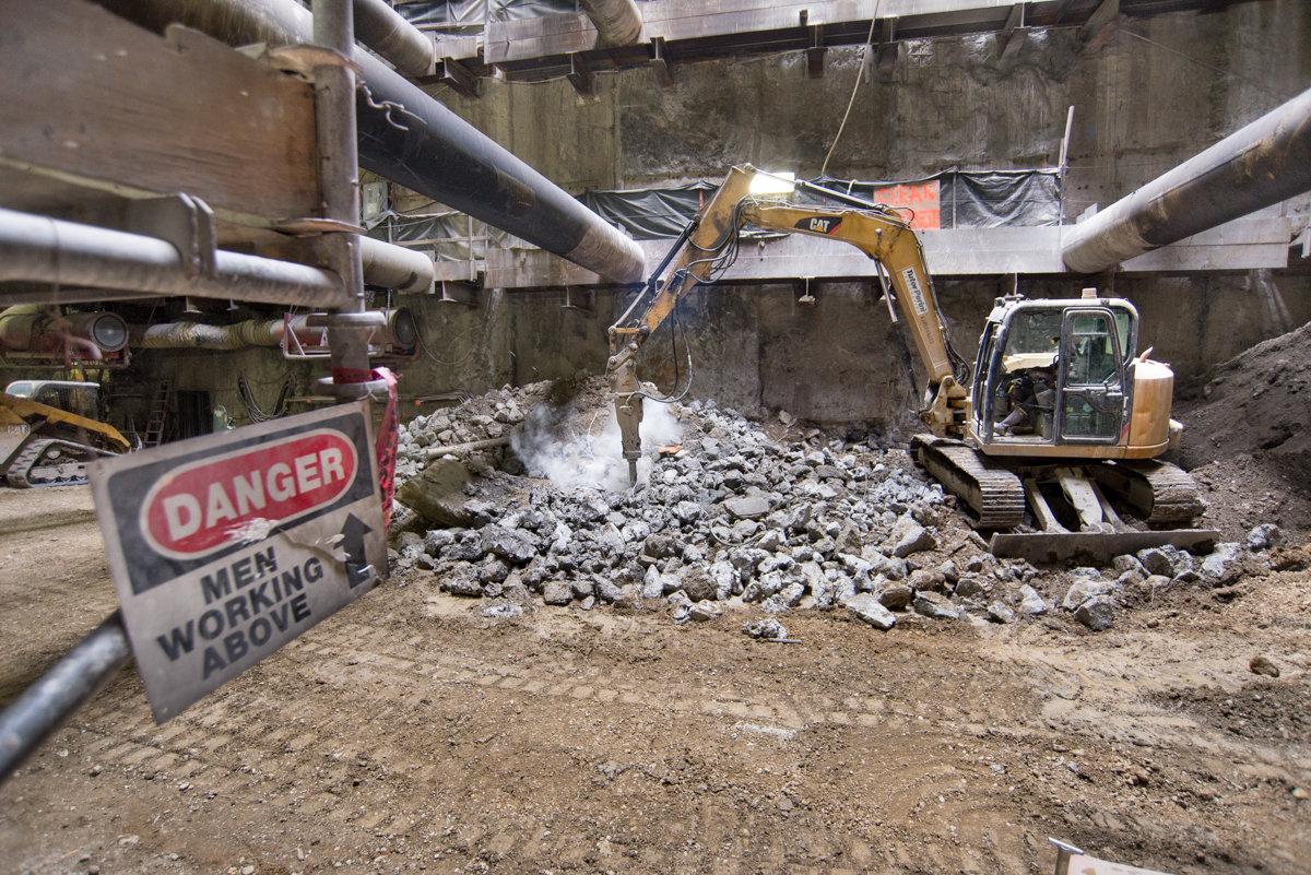 Excavator with jackhammer breaking apart concrete tunnel segments at underground transit station construction site.