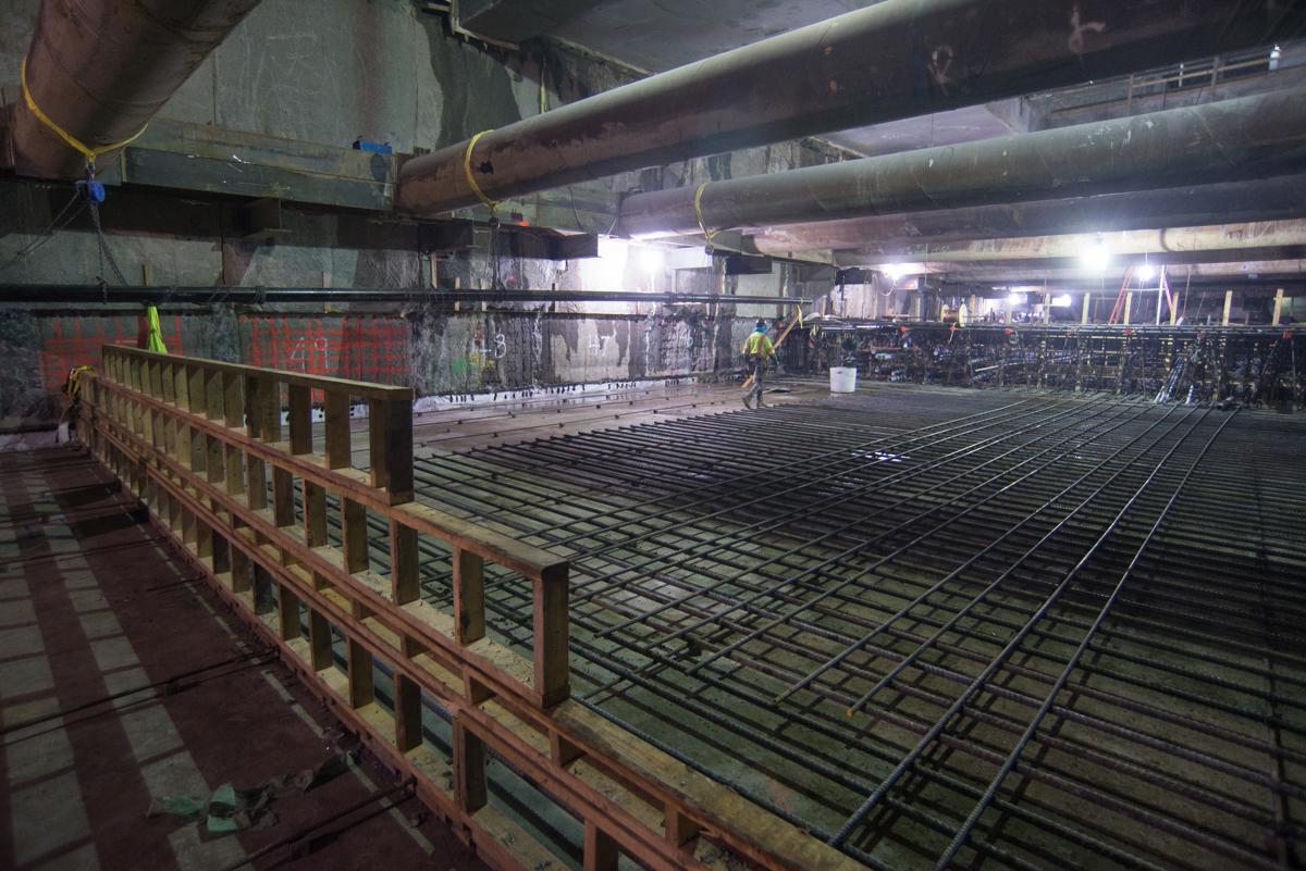 Industrial construction site with steel rebar reinforcement grid and temporary bracing under overhead pipes and beams.