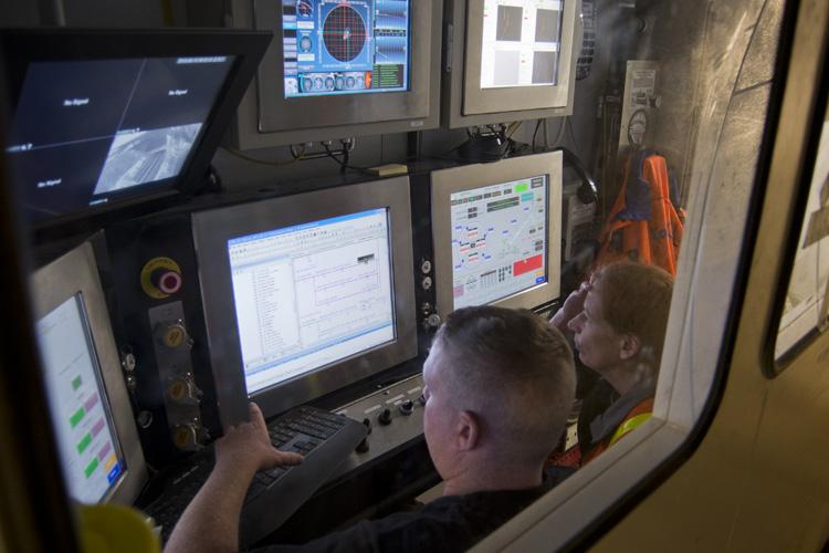 Two transit workers monitor multiple computer screens and displays in a tunnel boring machine control room.