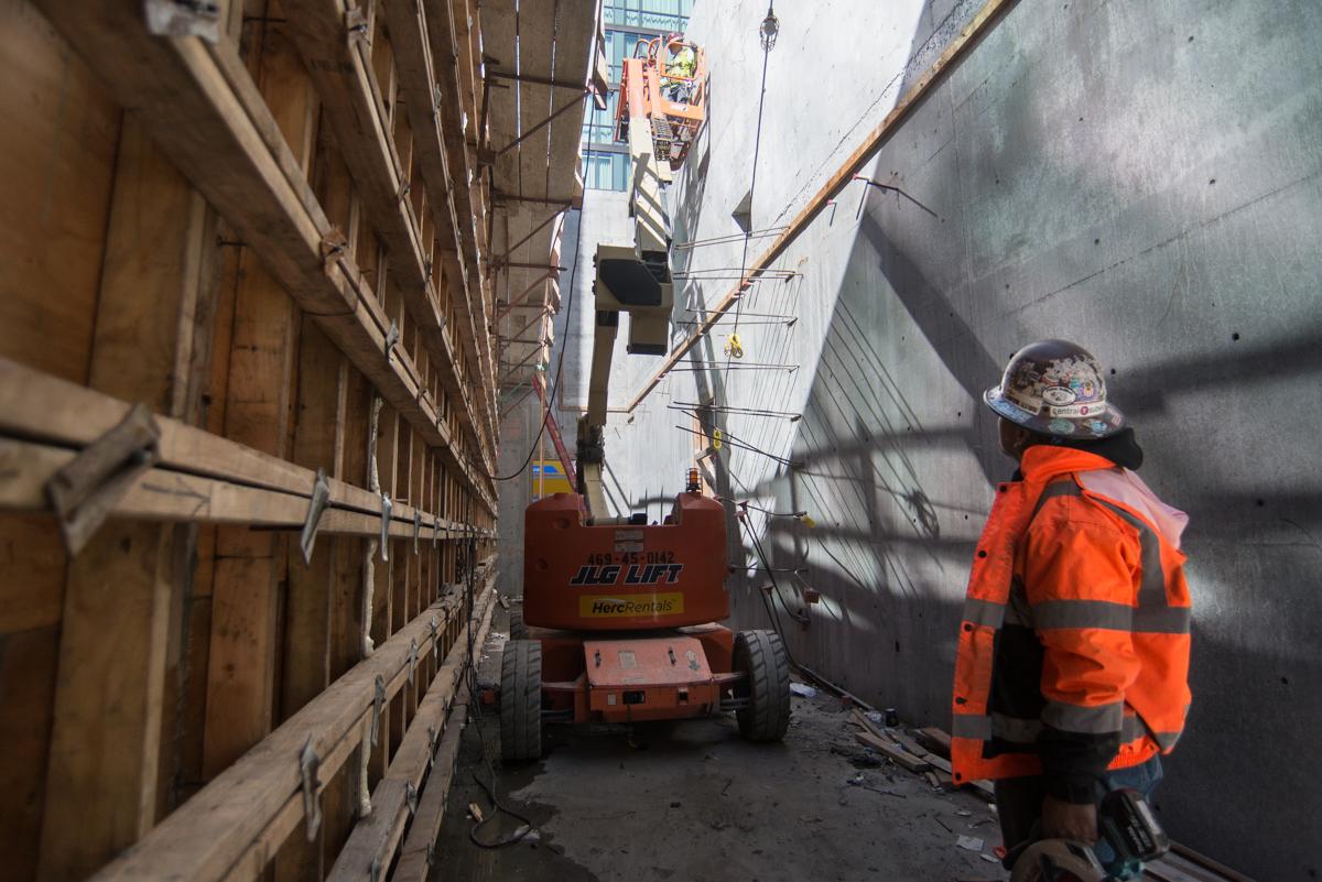 Worker in orange safety vest operating aerial lift between narrow concrete walls during construction or repair work.
