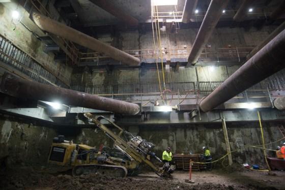 Construction equipment and workers inside underground Central Subway station tunnel with large pipes overhead.