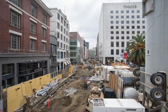 Construction site on Stockton Street with excavation equipment, yellow barriers, and surrounding downtown buildings.