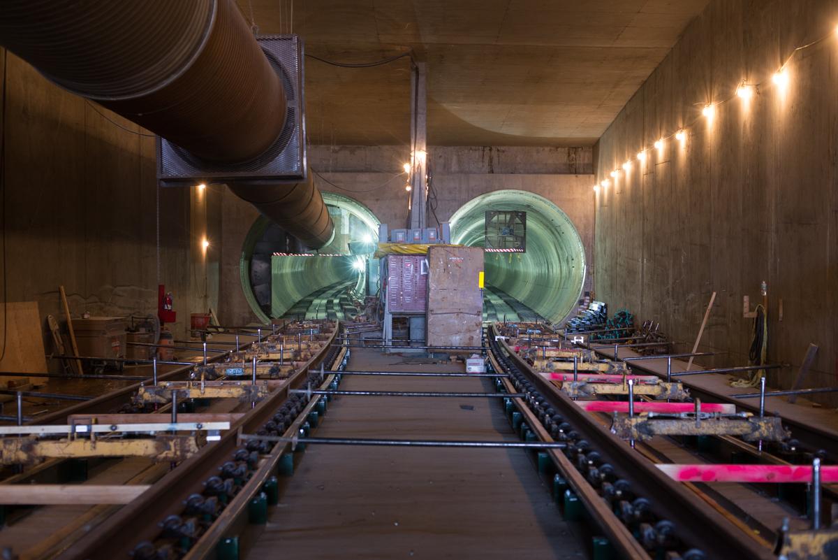 Tunnel interior with two large green tunnel boring machine heads visible, railroad tracks laid on the floor below, and construction equipment on either side.
