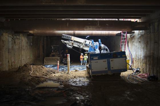 Construction equipment and workers operating machinery in an underground tunnel beneath a city street.