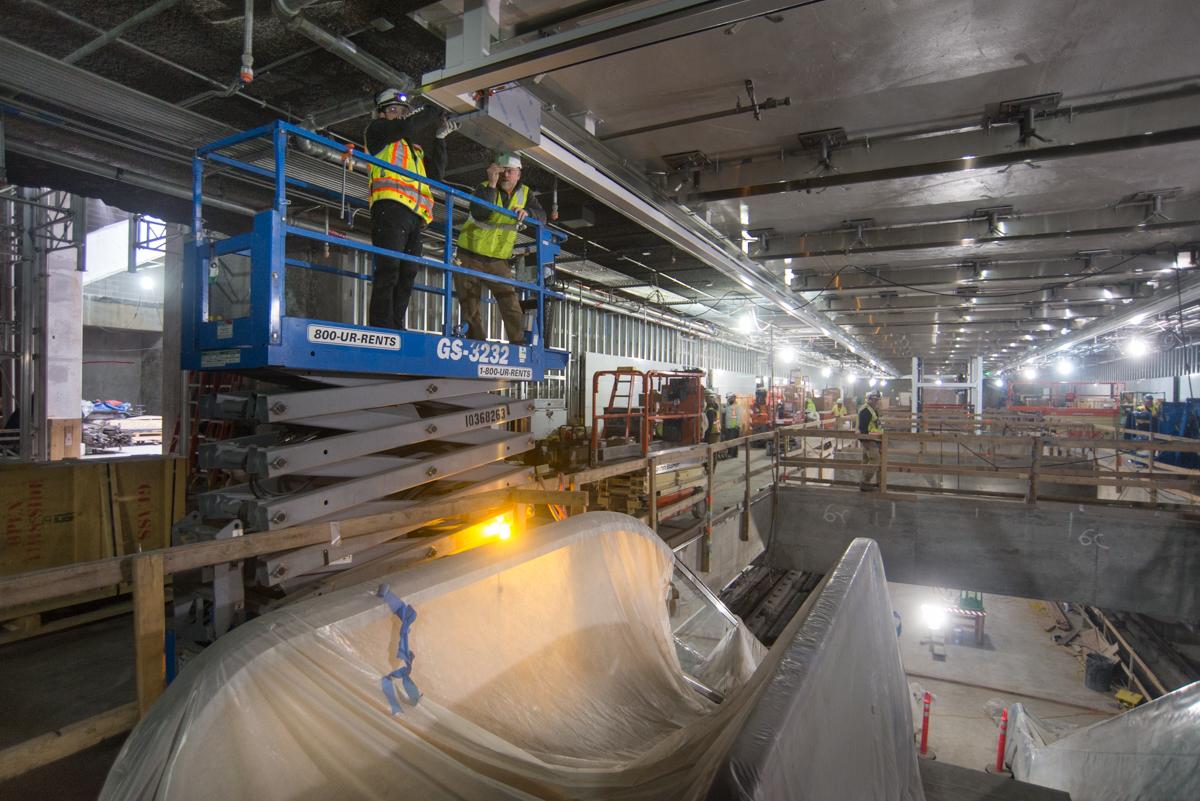 Workers on elevated lift platform installing ceiling section in transit station concourse during night shift.