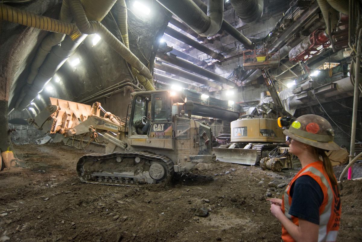 Construction worker in hard hat and safety vest observing CAT excavation equipment operating in underground tunnel with overhead lighting.