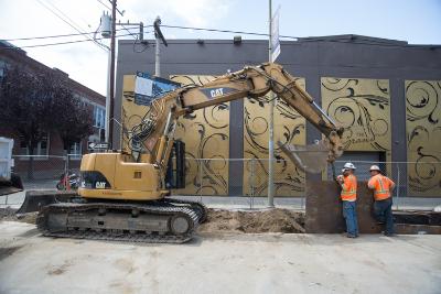 Yellow excavator and two workers in orange vests at construction site on urban street with mural-painted building.