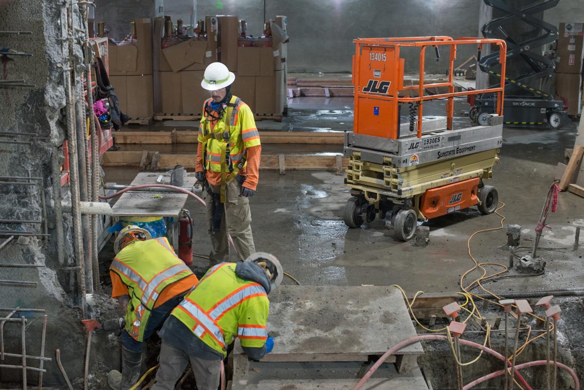 Construction workers in safety vests repair concrete around a slurry wall opening at a transit station using hand tools and equipment.