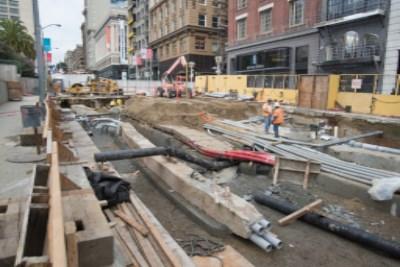 Construction site with exposed utilities and materials on concrete pads between urban buildings.