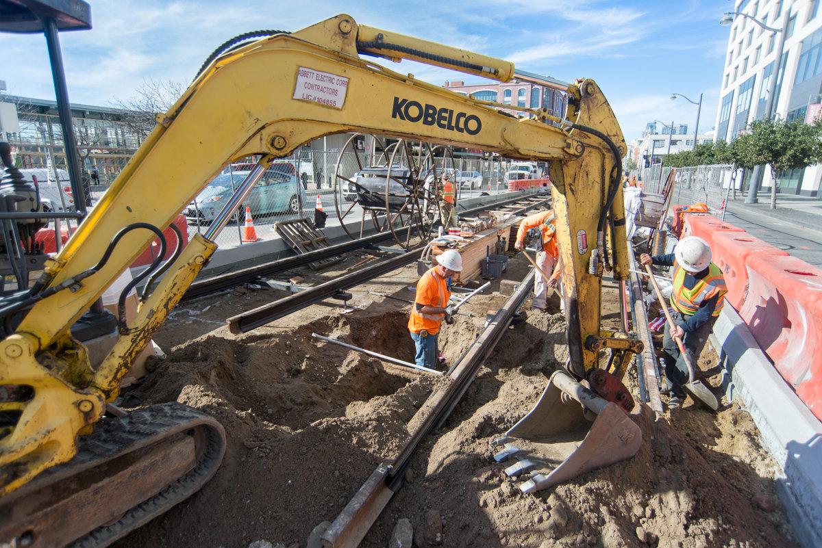 Yellow Kobelco excavator operates at urban construction site with workers laying transit rail tracks on city street.