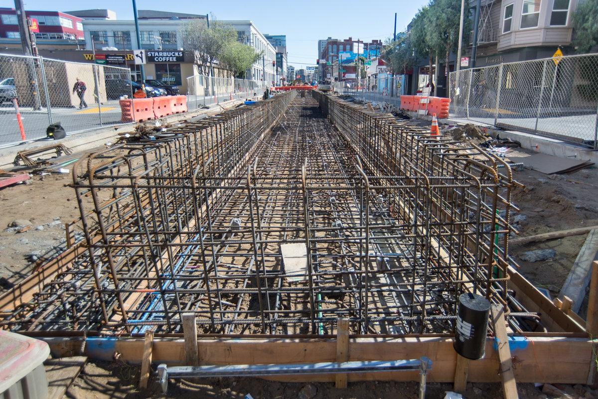 Construction site showing reinforced steel rebar framework for subway station foundation along urban street.