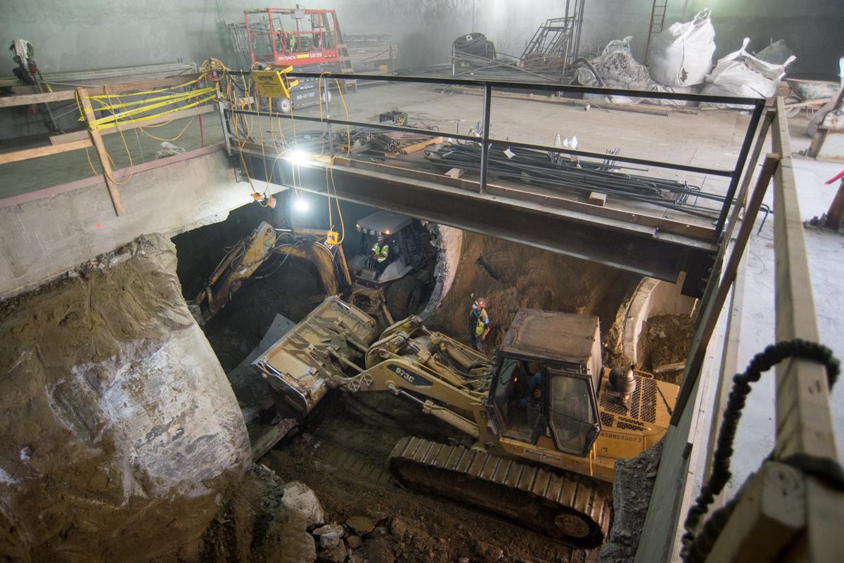 Workers excavate subway station box, breaking through twin tunnels with heavy equipment and lighting.
