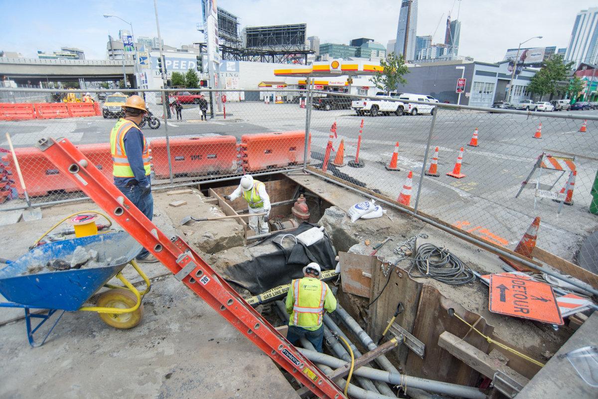 Construction workers in safety vests work in an excavated pit at 4th and Bryant Street, with orange barriers and traffic cones surrounding the site.