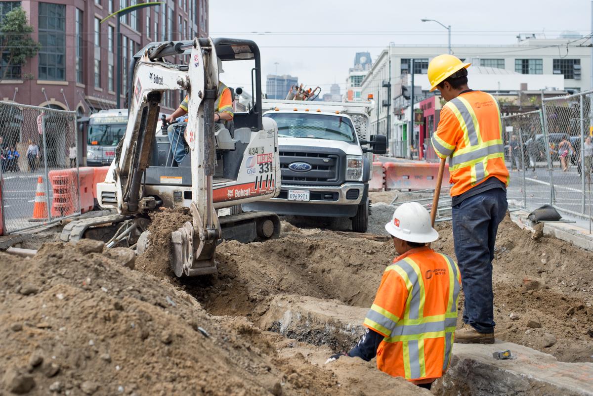 Construction workers operate an excavator to unearth utility infrastructure during Central Subway project work.