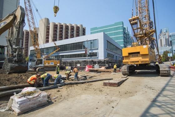 Construction site on 4th Street with excavators, workers, and building station walls amid downtown high-rises.