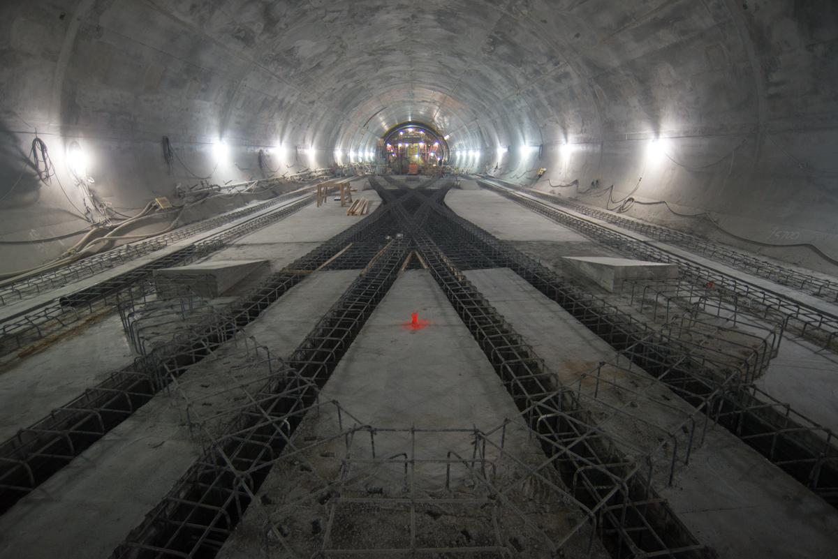 Underground tunnel with steel reinforcement framework and track crossover structure under construction, lit by work lights.