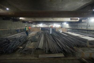 Underground transit station under construction with steel reinforcement bars stacked on floor and temporary support structures, worker visible.