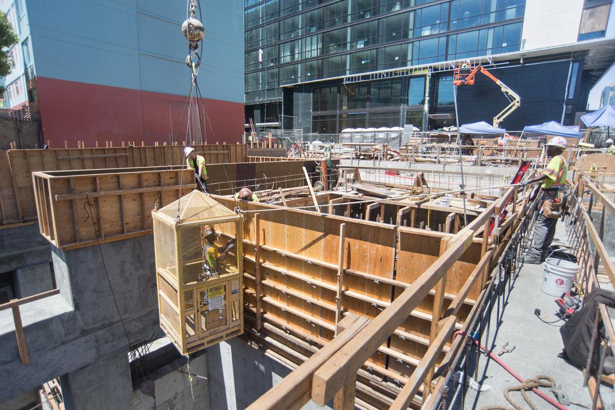 Construction workers build concrete forms in underground station headhouse with crane and safety equipment visible.