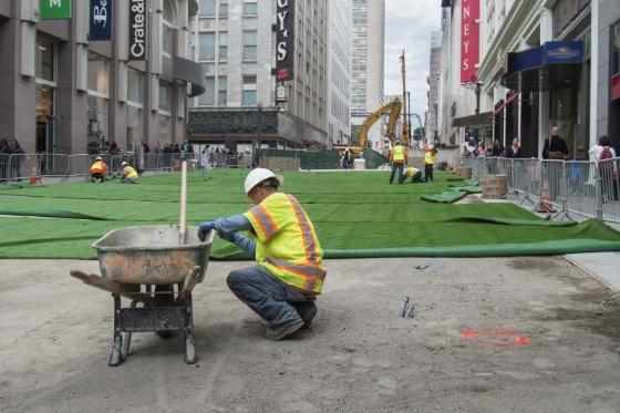 Worker in safety vest crouches beside fire pit on green carpet installation along urban street with tall buildings.