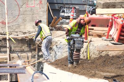 Construction workers operating pneumatic jackhammer on Stockton Street excavation site with heavy equipment nearby.